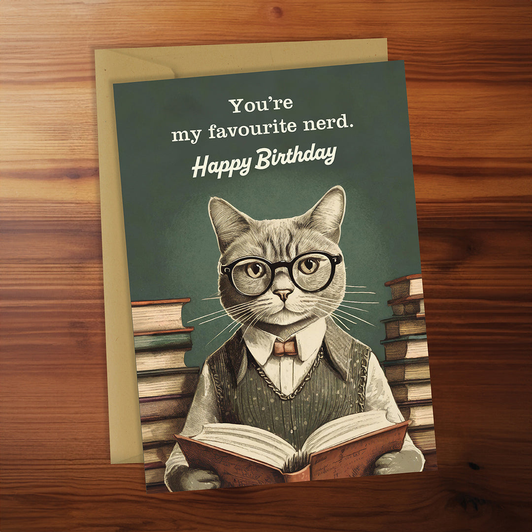Birthday card with a cat wearing glasses and reading a book, on a wooden surface.