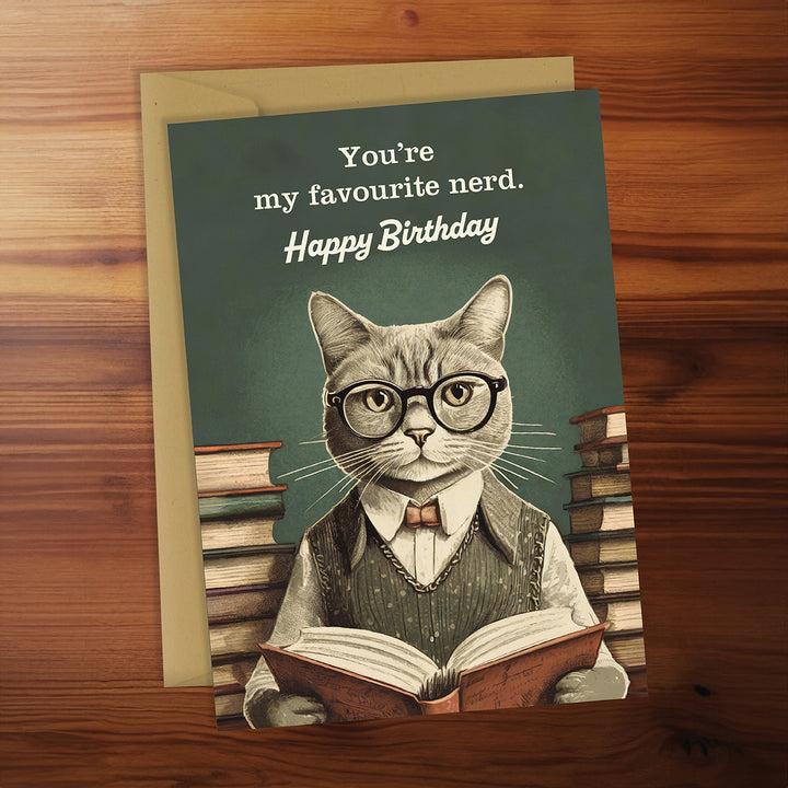 Birthday card with a cat wearing glasses and reading a book, on a wooden surface.