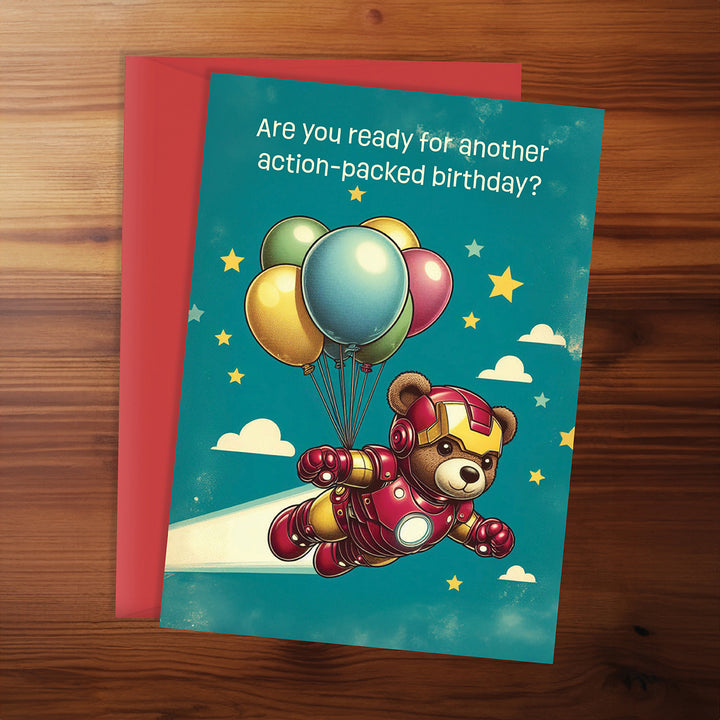 Birthday card with a teddy bear in superhero costume flying with balloons against a blue sky.